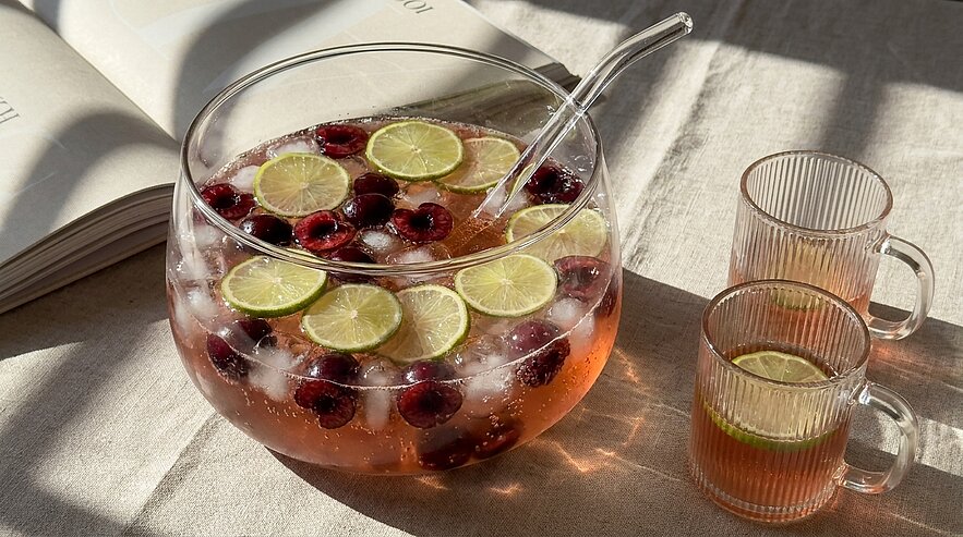 Glasbowle mit eisgekühltem Fruchtpunsch, Limettenscheiben und Kirschen, einem Glaslöffel und zwei gefüllten Gläsern daneben.