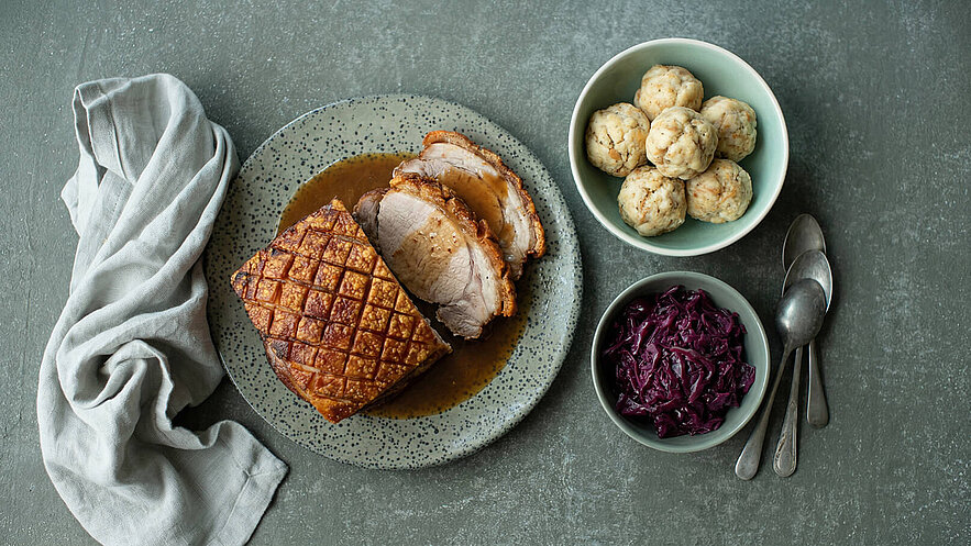 Ein Teller mit knusprigem Braten, in Scheiben geschnitten und in Soße serviert, steht neben zwei Schüsseln. Eine Schüssel enthält lila Rotkohl und die andere fünf knödelartige Beilagen. Ein beige Tuch liegt dekorativ im Bild.