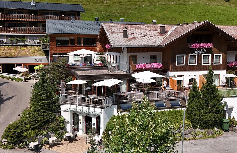 Das Bild zeigt ein rustikales Hotel mit mehreren Etagen, umgeben von grünen Wiesen. Auf dem Balkon und der Terrasse stehen Tische mit Sonnenschirmen. An der Fassade hängen Blumenkästen. Eine Straße führt zum Eingang des Hotels, der gut sichtbar ist.