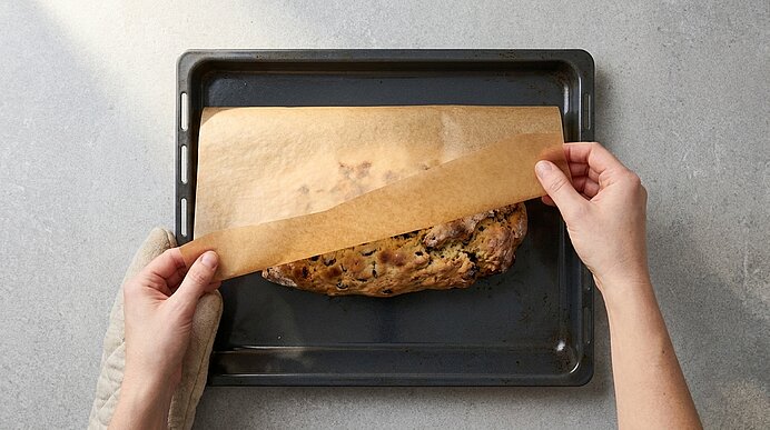Zwei Hände ziehen Backpapier von einem frisch gebackenen, goldbraunen Früchtebrot auf einem Backblech zurück.