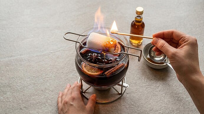 Eine Hand flambiert mit einem Brenner einen Marshmallow über einer Glasschale mit Glühwein, Zimtstangen, Sternanis und Orangenscheiben.