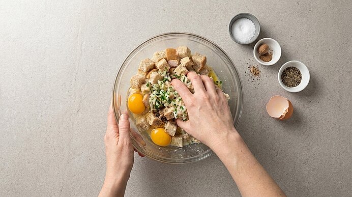 Zwei Hände mischen in einer Glasschüssel Eier, Brot‑würfel, Kräuter und Zwiebeln zu einer Füllung.