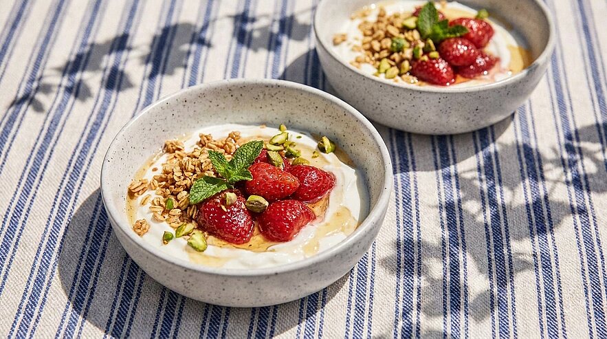 Zwei Keramikschalen mit Joghurt, garniert mit frischen Himbeeren, Granola, gehackten Pistazien, Honig und Minzblättern.