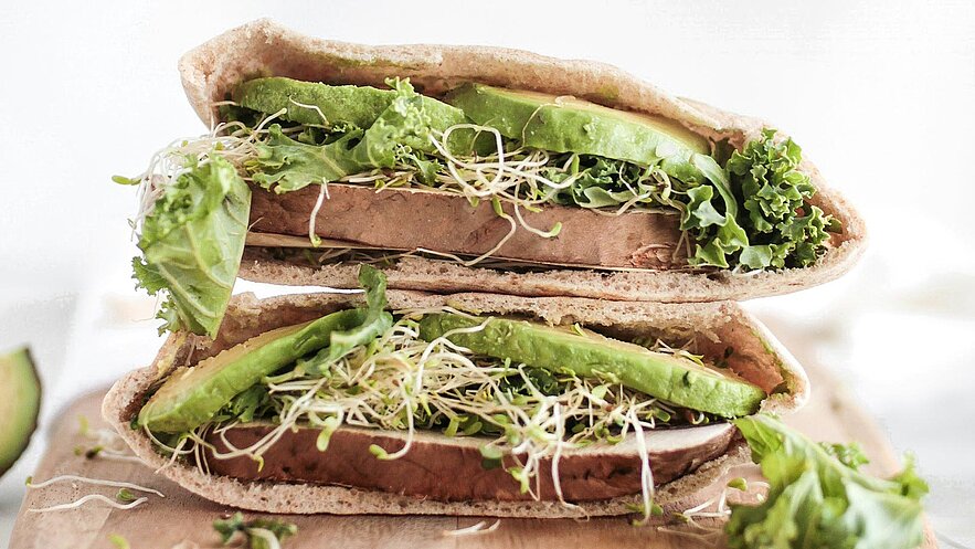 Ein aufgeschnittener Vollkorn-Pita-Brot mit frischem Gemüse. Darin befinden sich Schichten aus Salat, Avocado und Sprossen. Die Füllung ist üppig und grün, was einen gesunden Snack andeutet.
