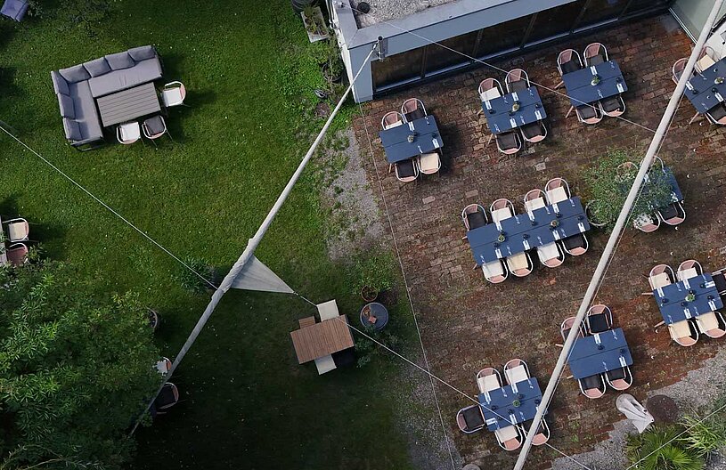 Eine Luftaufnahme eines Restaurantgarten mit mehreren Tischen und Stühlen. Auf der linken Seite sind gemütliche Sitzgelegenheiten, während rechts mehrere Tische auf einem gepflasterten Boden stehen. Ein Sonnensegel und Pflanzen schaffen eine einladende Atmosphäre.