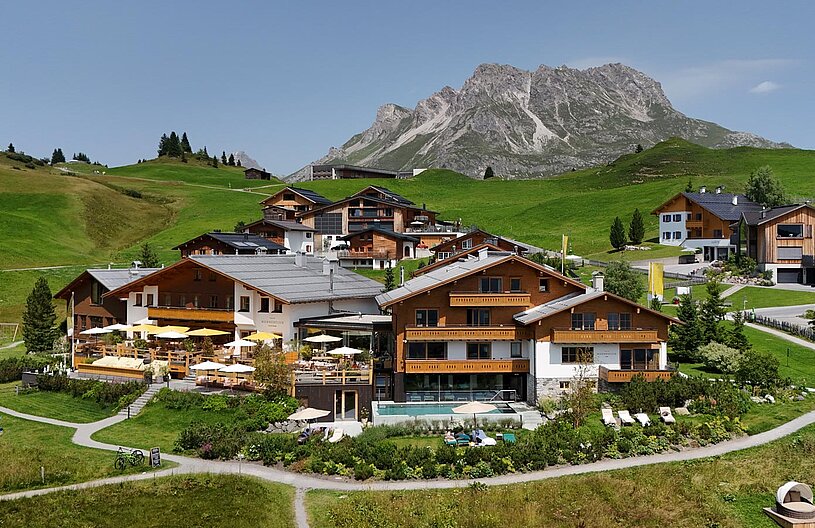 Eine malerische Alpenlandschaft mit mehreren modernen Chalets ist abgebildet. Die Gebäude sind umgeben von grünen Wiesen und Bäumen. Im Vordergrund ist eine Terrasse mit Sonnenschirmen und Liegen zu sehen, während im Hintergrund majestätische Berge aufragen.