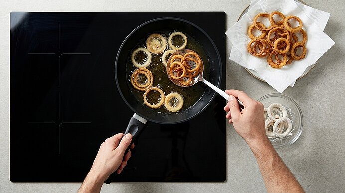 Zwei Hände heben frittierte Zwiebelringe mit einem Schaumlöffel aus heissem Öl in einer schwarzen Pfanne.