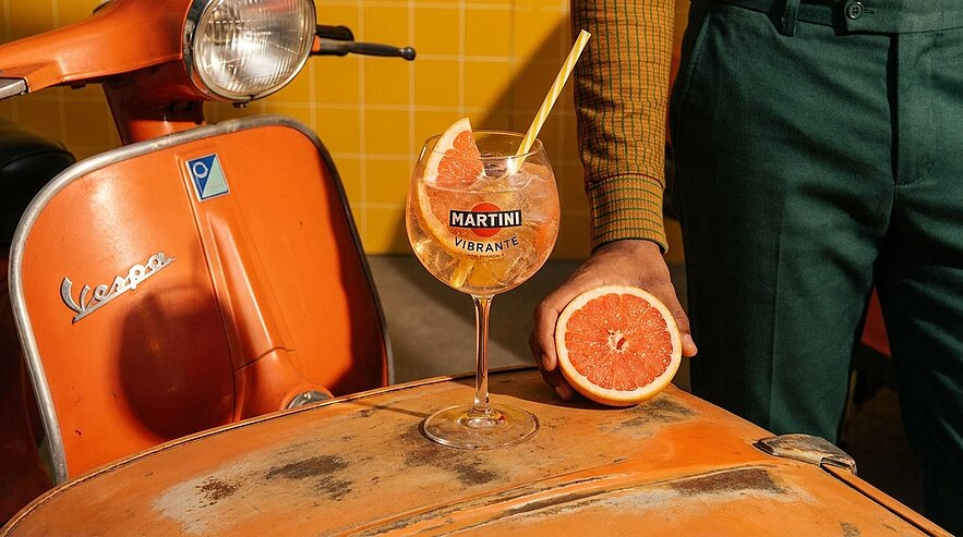 Orangefarbene Vespa im Vordergrund; auf ihrem Gepäckträger steht ein Martini-Glas mit Aufschrift "Martini Vibrante", gefüllt mit Grapefruit und Strohhalm. Daneben hält eine Person eine halbierte Grapefruit.