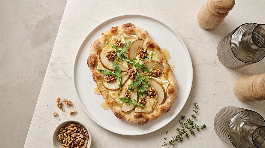 Eine mit dünnen Birnenscheiben, Walnüssen und frischem Rucola belegte Pizza auf einem weißen Teller. Neben dem Teller befindet sich eine kleine Schale mit Walnüssen. Der Hintergrund ist ein heller, einfarbiger Tisch.