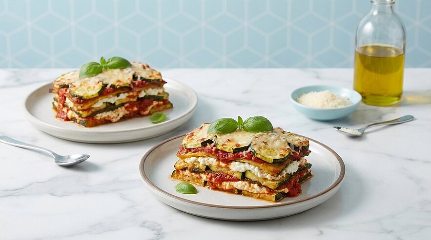 Zwei Portionen geschichtete Gemüse-Lasagne mit Zucchinischeiben, Tomatensauce und geschmolzenem Käse, jeweils mit frischen Basilikumblättern garniert, auf Tellern serviert.
