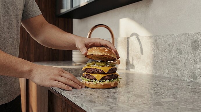 Hände einer Person platzieren einen großen, doppelt belegten Cheeseburger mit zwei Rindfleisch‑Patties, geschmolzenem Käse, Salat und Gurkenscheiben auf einer Küchenarbeitsplatte.