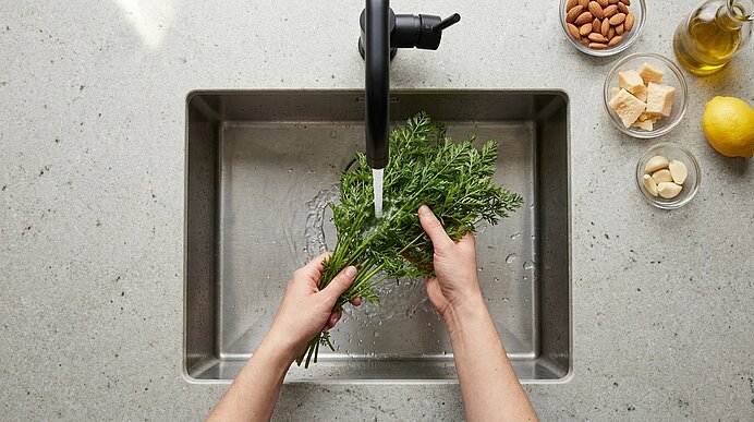 Draufsicht: Hände spülen frische Kräuter unter fließendem Wasser am Wasserhahn in einem Küchenspülbecken.