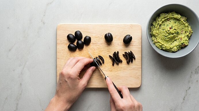 Zwei Hände schneiden schwarze Oliven auf einem Holzbrett in Streifen. Rechts steht eine Schale mit Guacamole.