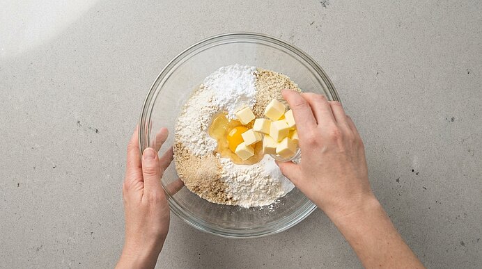 Zwei Hände fügen gewürfelte Butter und ein Ei zu Mehl und trockenen Zutaten in einer Glasschüssel hinzu.