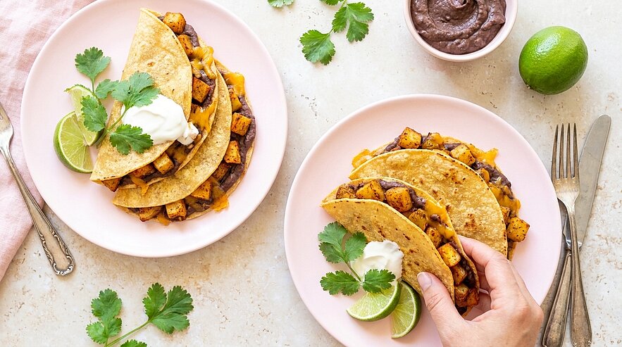 Zwei Teller mit gefalteten Tortillas, gefüllt mit gewürzten Kartoffelwürfeln und Bohnen, garniert mit saurer Sahne, Koriander und Limetten; eine Hand hält einen Taco.