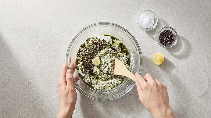 Zwei Hände verrühren Topfen, Kürbiskerne, Kernöl und Knoblauch mit einem Holzspatel in einer Glasschüssel.
