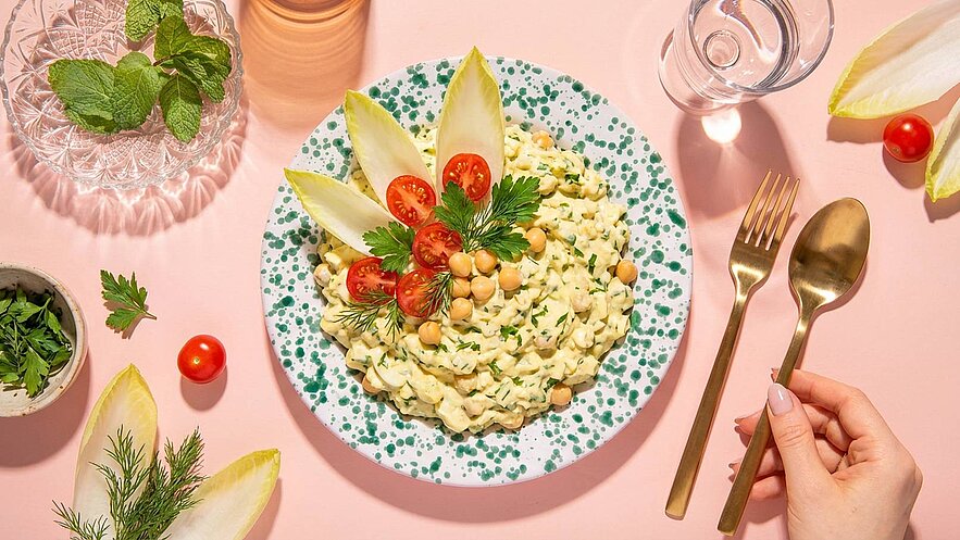 Ein Teller mit cremigem Salat aus Pasta, Kichererbsen und frischen Kräutern. Oben auf sind Cherry-Tomaten und Endiviensalat dekoriert. Daneben liegen eine Gabel und ein Löffel aus Gold. Der Hintergrund ist in zartem Rosa gehalten.