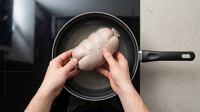 Zwei Hände legen eine mit Küchengarn gebundene Wurst- oder Fleischrolle in eine Pfanne mit Wasser auf einem Herd.