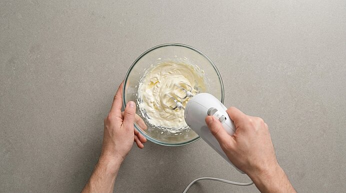 Zwei Hände schlagen Butter, Sauerrahm und Salz in einer Glasschüssel mit einem Handrührgerät auf.