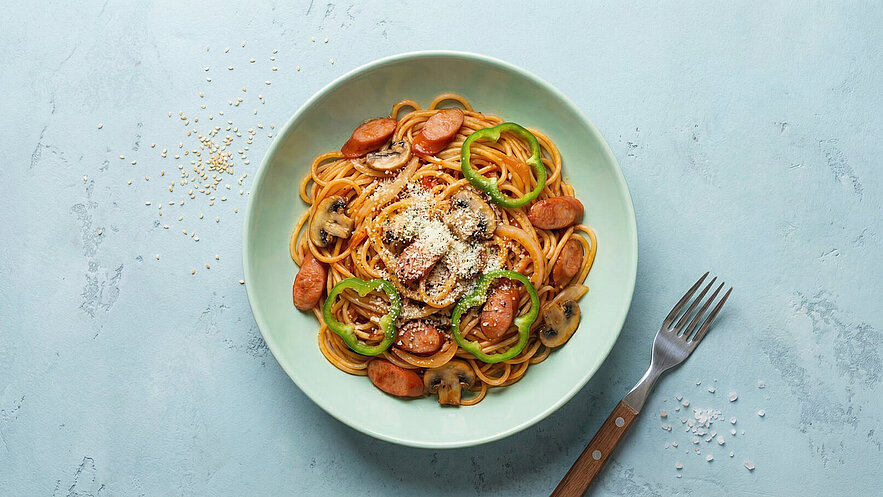 Ein Teller Spaghetti mit Saucenwurst, grünen Paprikastreifen und Champignons, garniert mit Parmesan. Der Teller ist mintgrün und befindet sich auf einer hellen Oberfläche, daneben liegt eine Gabel.