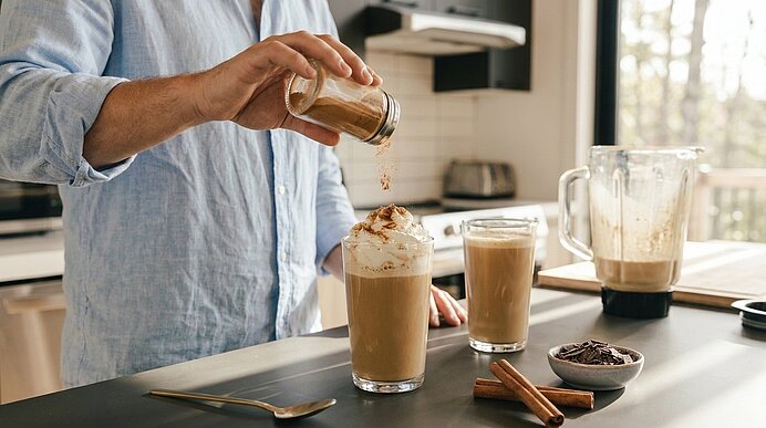 Person in hellem Hemd bestreut den Milchschaum eines Latteglases mit Zimt; zwei Lattegläser, Mixer, Zimtstangen und Schokoraspeln auf der Küchentheke.