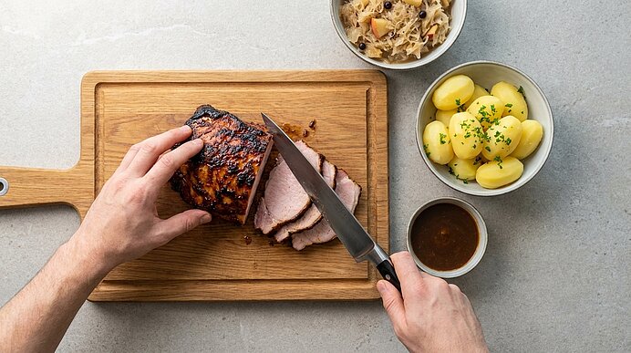 Hände schneiden glasierten Braten auf einem Holzbrett; daneben Schalen mit Petersilienkartoffeln, Sauerkraut und Bratensoße.