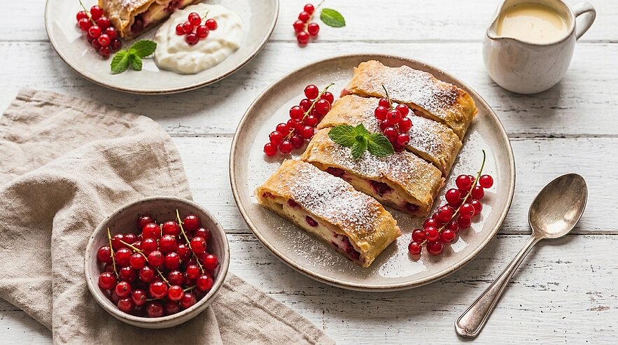 Drei Scheiben Strudel mit roten Johannisbeeren auf einem Teller, mit Puderzucker bestäubt und mit Minzblatt garniert; daneben eine Schale frischer Johannisbeeren und eine kleine Saucière mit Vanillesauce.