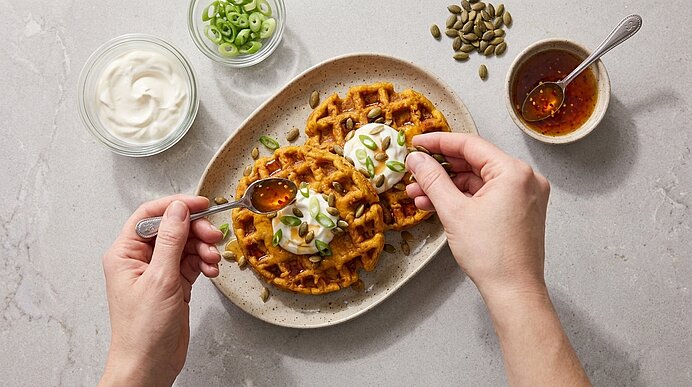 Zwei orangefarbene Waffeln auf einem Teller, garniert mit Sauerrahm, Frühlingszwiebeln und Kürbiskernen; eine Hand beträufelt sie mit Honig oder Sirup.