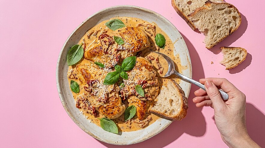 Teller mit gebratenen Hähnchenfilets in cremiger Tomaten‑Basilikum‑Sauce, garniert mit Basilikum und geriebenem Käse; daneben rustikales Brot, eine Hand taucht ein Brotstück in die Sauce.