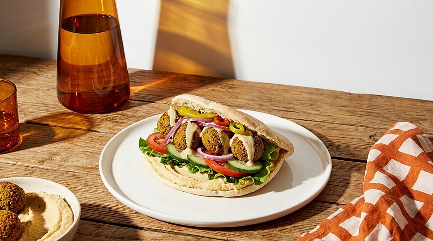 Pitabrot gefüllt mit Falafelbällchen, Salat, Tomatenscheiben, roten Zwiebeln und Sauce, angerichtet auf einem weißen Teller.