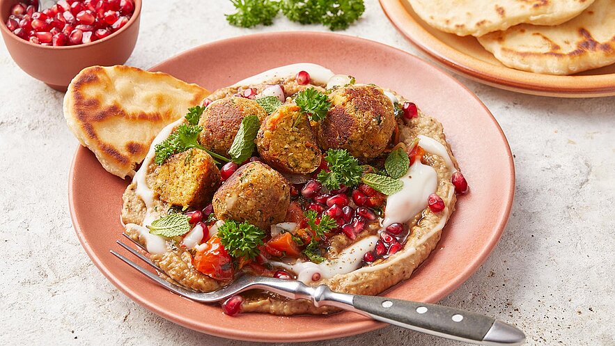 Ein Teller mit Falafel, garniert mit frischem Gemüse, Joghurt und Granatapfelkernen, auf einer Basis aus pürierten Hülsenfrüchten. Daneben liegt ein Stück Fladenbrot. Im Hintergrund sind weitere Fladenbrote sichtbar.