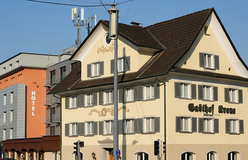 Eine Ansicht zweier Hotels im Stadtbild. Links steht ein älteres, gelb gestrichenes Gebäude mit vielen Fenstern und dem Namen "Gasthof Arne". Rechts ist ein moderner, grauer Bau mit der Aufschrift "HOTEL" in großen, weißen Buchstaben. Im Hintergrund ein blauer Himmel.