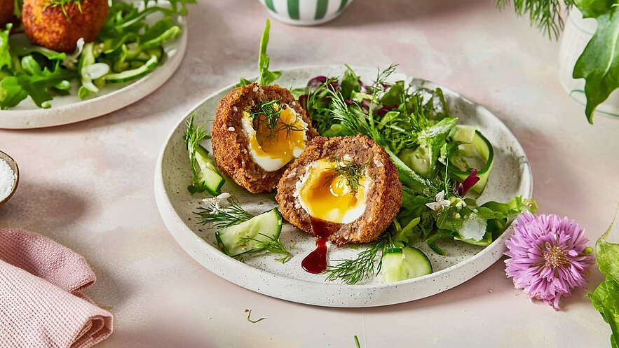 Auf dem Bild sind zwei aufgeschnittene, frittierte Eierbällchen mit flüssigem Eigelb zu sehen, serviert auf einem Teller mit frischem Salat und Gurkenscheiben. Das Gericht ist mit frischen Kräutern garniert.