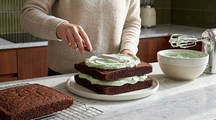 Hände einer Person bestreichen gestapelte Schokoladenkuchen mit hellgrüner Buttercreme; Schüssel mit Creme und Handmixer auf der Arbeitsfläche daneben.