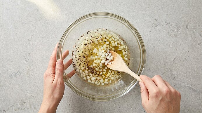 Zwei Hände rühren mit einem Holzlöffel gewürfelte Zwiebeln in einer Glasschüssel mit Öl und Kräutern.