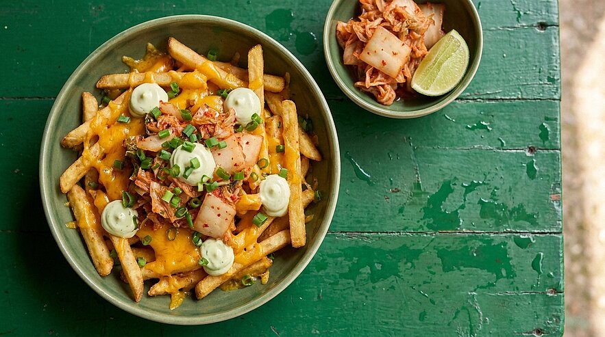 Schale mit überbackenen Pommes frites: geschmolzener Käse, kimchi-Stücke, grünen Creme‑Tupfen und gehackten Frühlingszwiebeln; daneben kleine Schüssel mit zusätzlichem Kimchi und Limettenviertel.