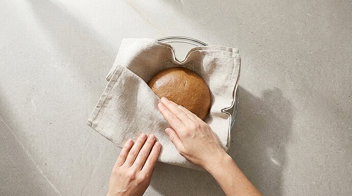 Von oben aufgenommen: zwei Hände legen ein rundes, braunes Brot in einen mit Leinen ausgelegten Brotkorb.