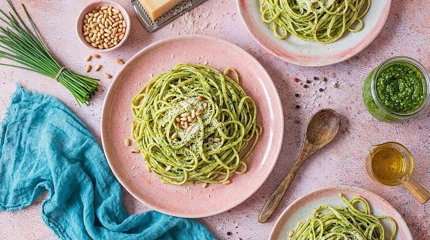 Ein gedeckter Tisch mit zwei Tellern Pasta, die mit grünem Pesto, Pinienkernen und geriebenem Käse angerichtet sind. Daneben befinden sich ein Glas Pesto, Olivenöl und eine Holzlöffel. Ein frischer blauer Stoff und frische Schnittlauchzweige runden das Bild ab.