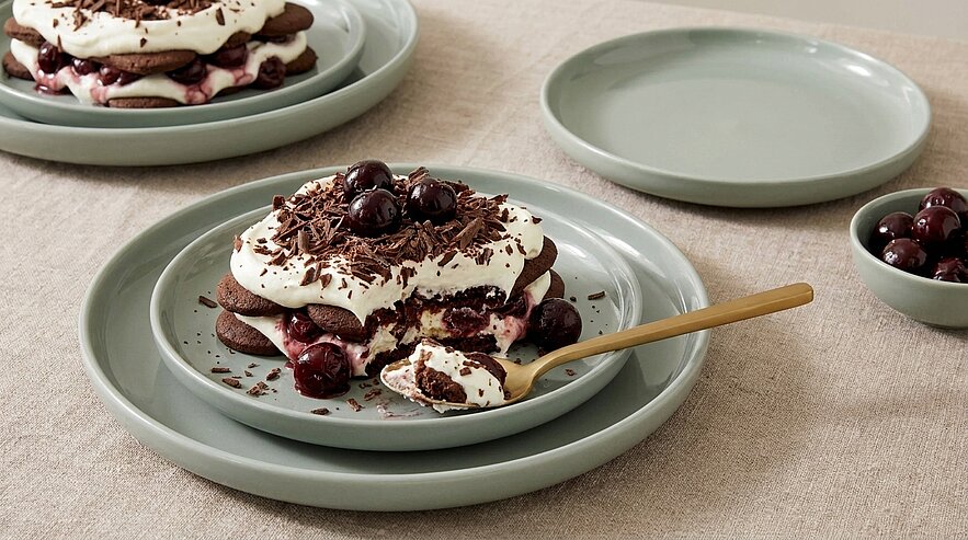 Schichtdessert aus Schokoladenböden mit Schlagsahne, ganzen Kirschen und Schokoraspeln auf einem Teller, eine Portion mit Löffel.