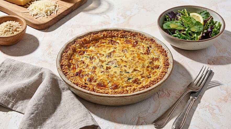 Goldbraune Quiche in runder Backform auf hellem Tisch, daneben eine Schüssel mit grünem Salat sowie Gabeln und eine Serviette.