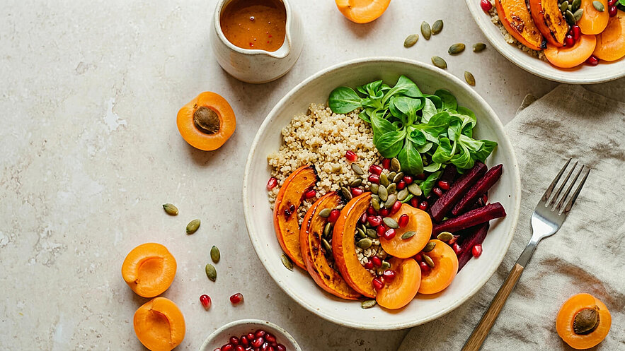 Eine Schüssel mit Quinoa, gebackenem Kürbis, frischem Spinat, roten Bete-Streifen und Aprikosenstücken. Darüber sind Kürbiskerne und Granatapfelkerne verteilt. Neben der Schüssel befindet sich eine kleine Kanne mit einer Soße.