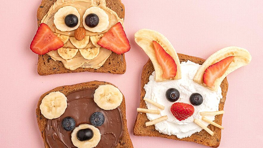 Drei Scheiben Brot, dekoriert als Tiergesichter: Oben ein Hase mit Quark, Bananen und Erdbeeren. Unten ein Bär mit Nutella, Bananen und Heidelbeeren. Ein dritter Toast zeigt ein Schaf mit Erdnussbutter, Bananen und Erdbeeren. Farbenfroh und einladend.