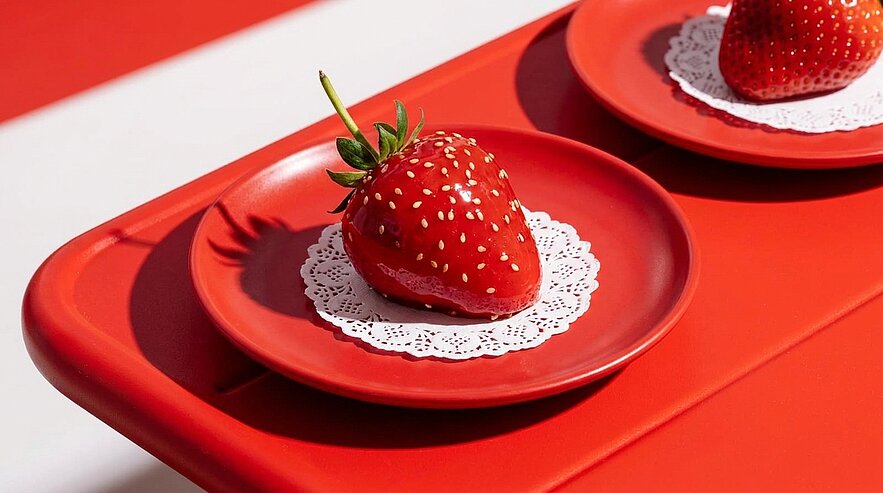 Zwei große glänzende Erdbeeren liegen jeweils auf kleinen roten Tellern auf weißen Spitzendeckchen, in einer reduzierten roten, minimalistischen Präsentation.