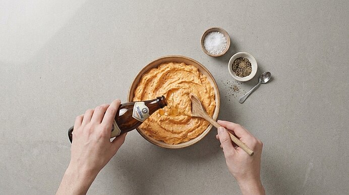 Zwei Hände rühren in einer Schüssel eine cremige, orangefarbene Masse mit einem Holzlöffel, während aus einer Bierflasche Flüssigkeit zugegeben wird; daneben kleine Schälchen mit Salz und Pfeffer.