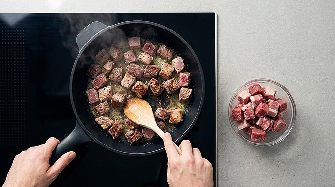 Draufsicht: Hände rühren mit einem Holzlöffel angebratene Rindfleischwürfel in einer Pfanne auf dem Herd; daneben eine Schüssel mit rohen Fleischwürfeln.