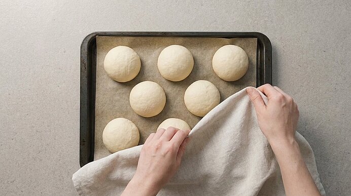 Zwei Hände decken geformte Pizzabrötchen auf einem Backblech mit einem hellen Tuch ab.