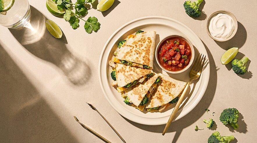 Teller mit vier goldbraun gebratenen Quesadilla-Dreiecken, gefüllt mit Käse und grünen Kräutern, daneben kleine Schale Tomatensalsa und Limettenspalten.