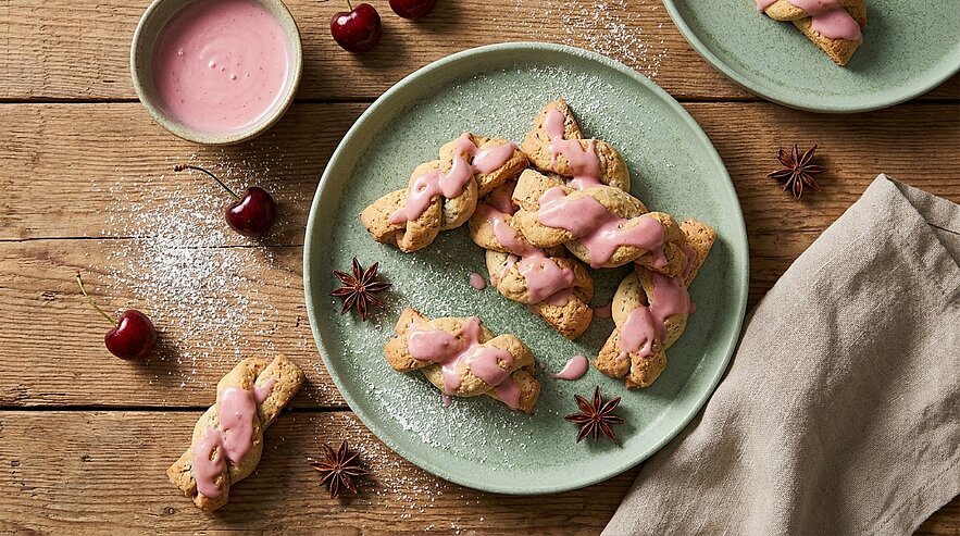 Ein Teller mit geflochtenen Keksen, die mit einer rosa Glasur dekoriert sind. Neben dem Teller befinden sich Kirschen und eine kleine Schüssel mit rosa Sauce. Der Untergrund ist aus Holz, und es streuen einige Zucker- oder Mehlspuren auf der Oberfläche.