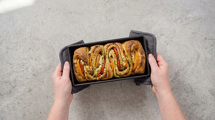 Zwei Hände mit grauen Topflappen halten eine Kastenform mit frisch gebackenem, goldbraunem Faltenbrot.
