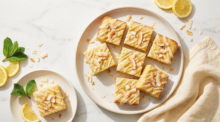 Großer Teller mit mehreren quadratischen Zitronenkuchenstücken, mit gerösteten Kokosflocken bestreut; daneben ein einzelnes Stück auf kleinem Teller, Zitronenscheiben und Minzblatt.